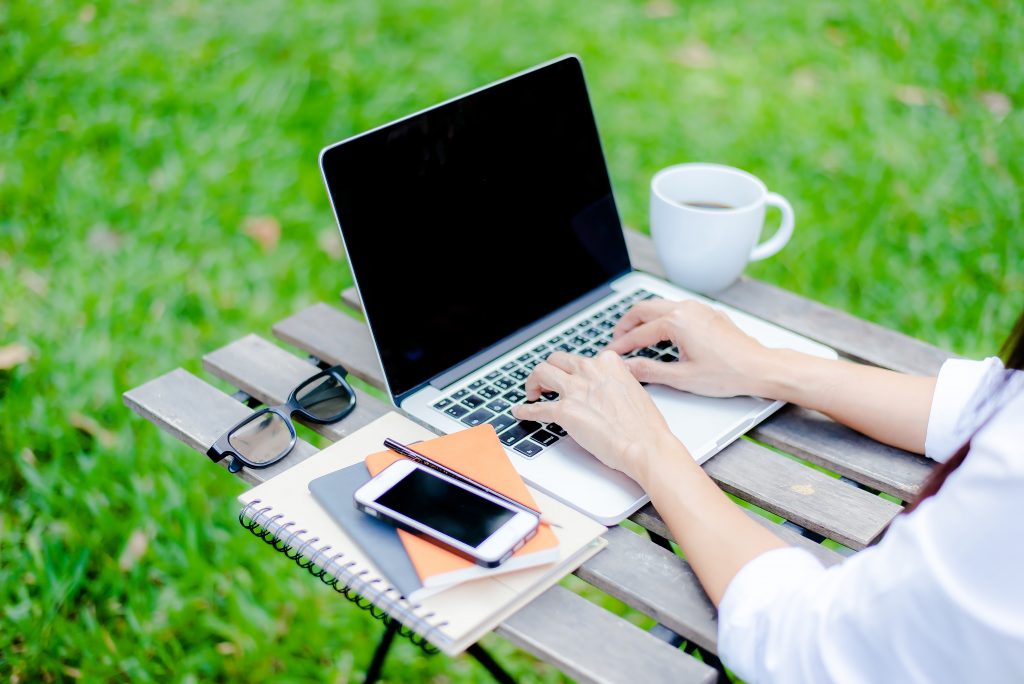 Freelance work. Casual dressed man sitting at wooden desk inside garden working on computer pointing with color pen electronic gadgets dropped around on table side view